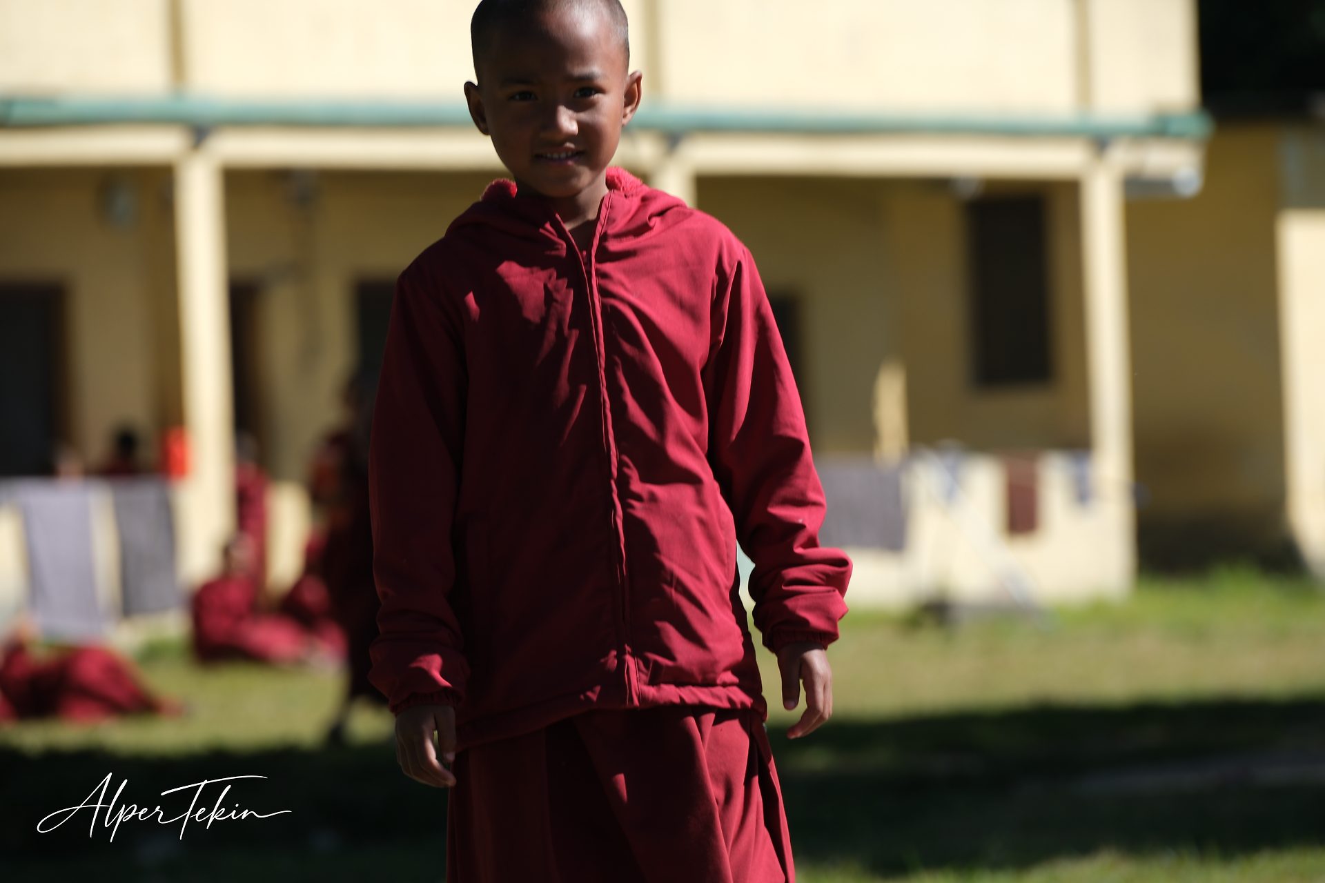 A young monk in a maroon hooded jacket and robe looks at the camera on a sunlit lawn, with a yellow building and blurred seated figures behind.