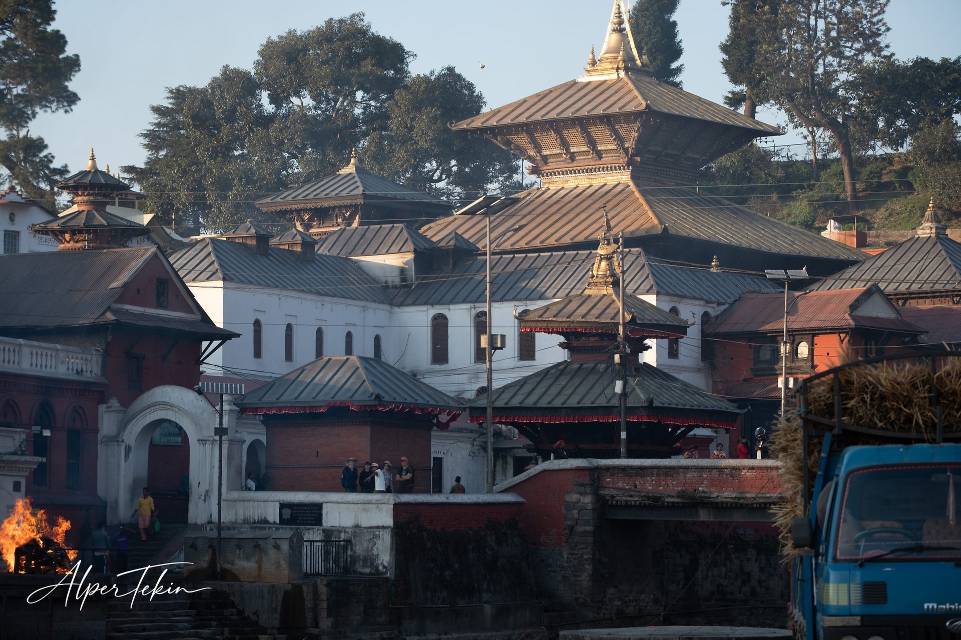 Multi-tiered pagoda temple with a gilded pinnacle and copper roofs among trees; a fire burns at lower left, a blue truck loaded with hay at right.