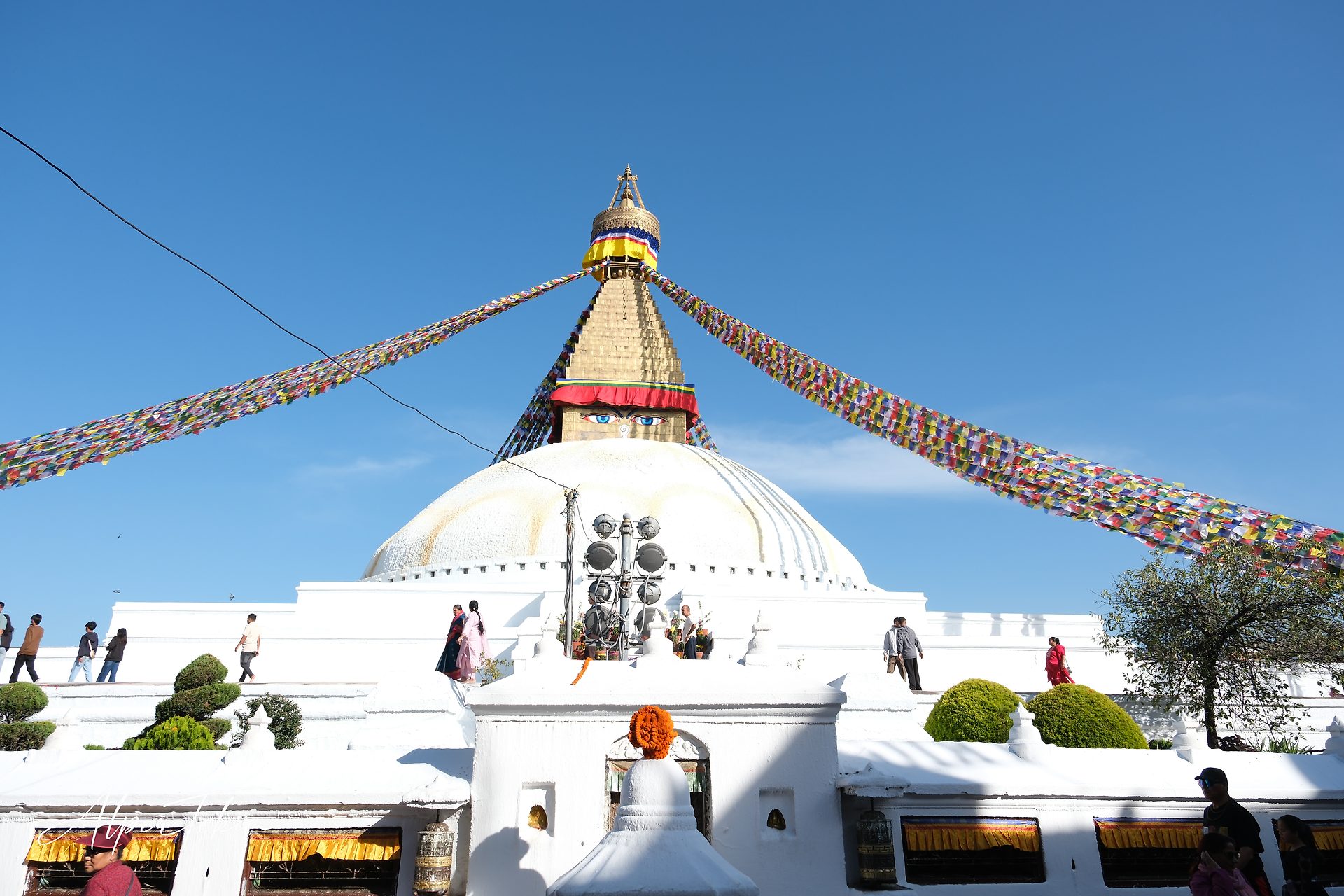 White-domed Boudhanath stupa with Buddha eyes on its golden tower, colorful prayer flags stretching to both sides, a clear blue sky and people walking around.