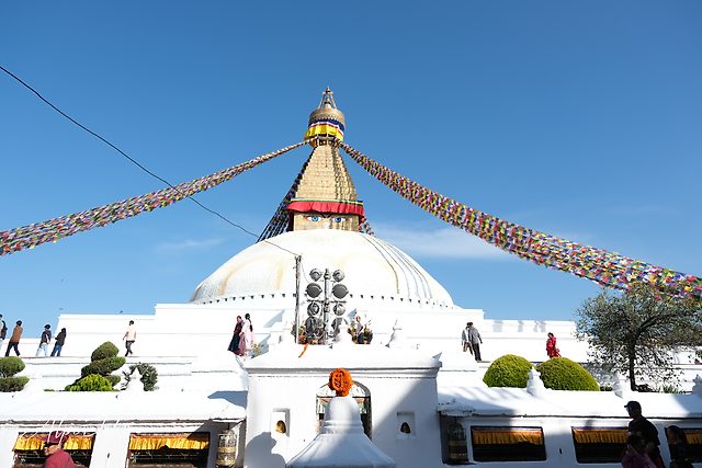 White-domed Boudhanath stupa with Buddha eyes on its golden tower, colorful prayer flags stretching to both sides, a clear blue sky and people walking around.