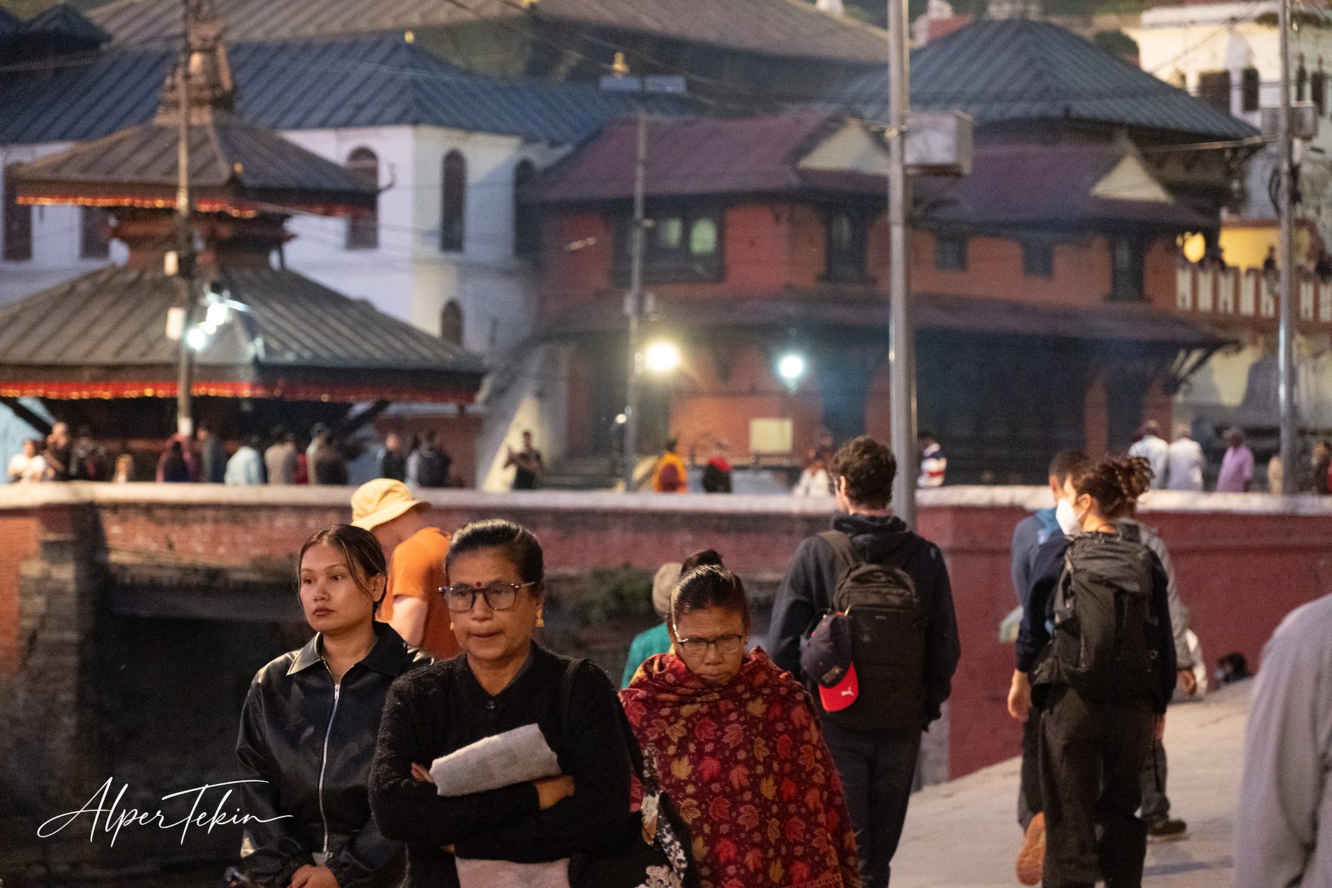 People walking along a low brick wall at dusk; two women with glasses in front, tiered-roof brick buildings and lit lamps behind them.
