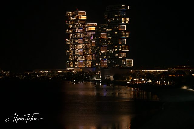 Two lit towers of offset, stacked boxes against a black night sky, with calm dark water in front reflecting their orange light.