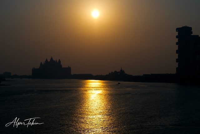 A bright sun in a hazy orange sky, a golden path of light on dark water, and silhouettes of a turreted building and a tall tower on the horizon.