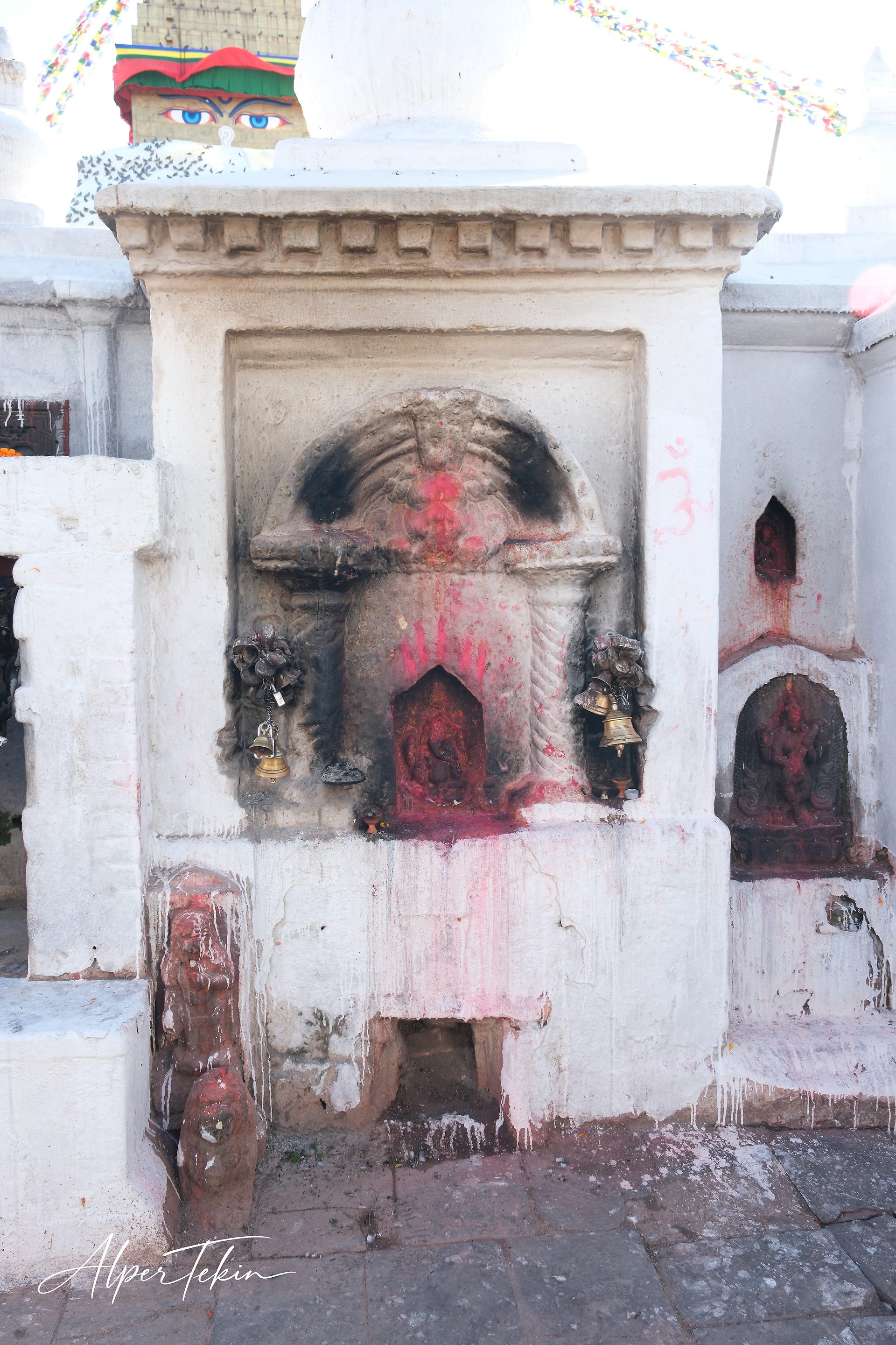 A soot-darkened niche on a whitewashed wall, smeared with red powder and flanked by brass bells, with the painted Buddha eyes and prayer flags behind.