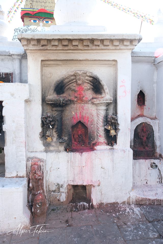 A soot-darkened niche on a whitewashed wall, smeared with red powder and flanked by brass bells, with the painted Buddha eyes and prayer flags behind.