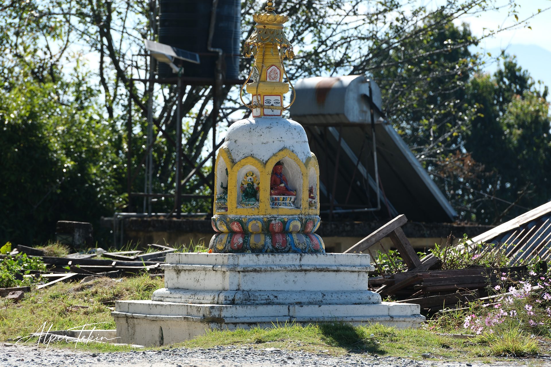 A small Buddhist stupa with yellow arches and a gilded spire on a white base, set against a water tank, solar heater, trees and scattered planks.