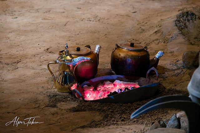Two copper kettles and a golden Arabic coffee pot sit on sandy ground beside a metal pan of glowing red embers.