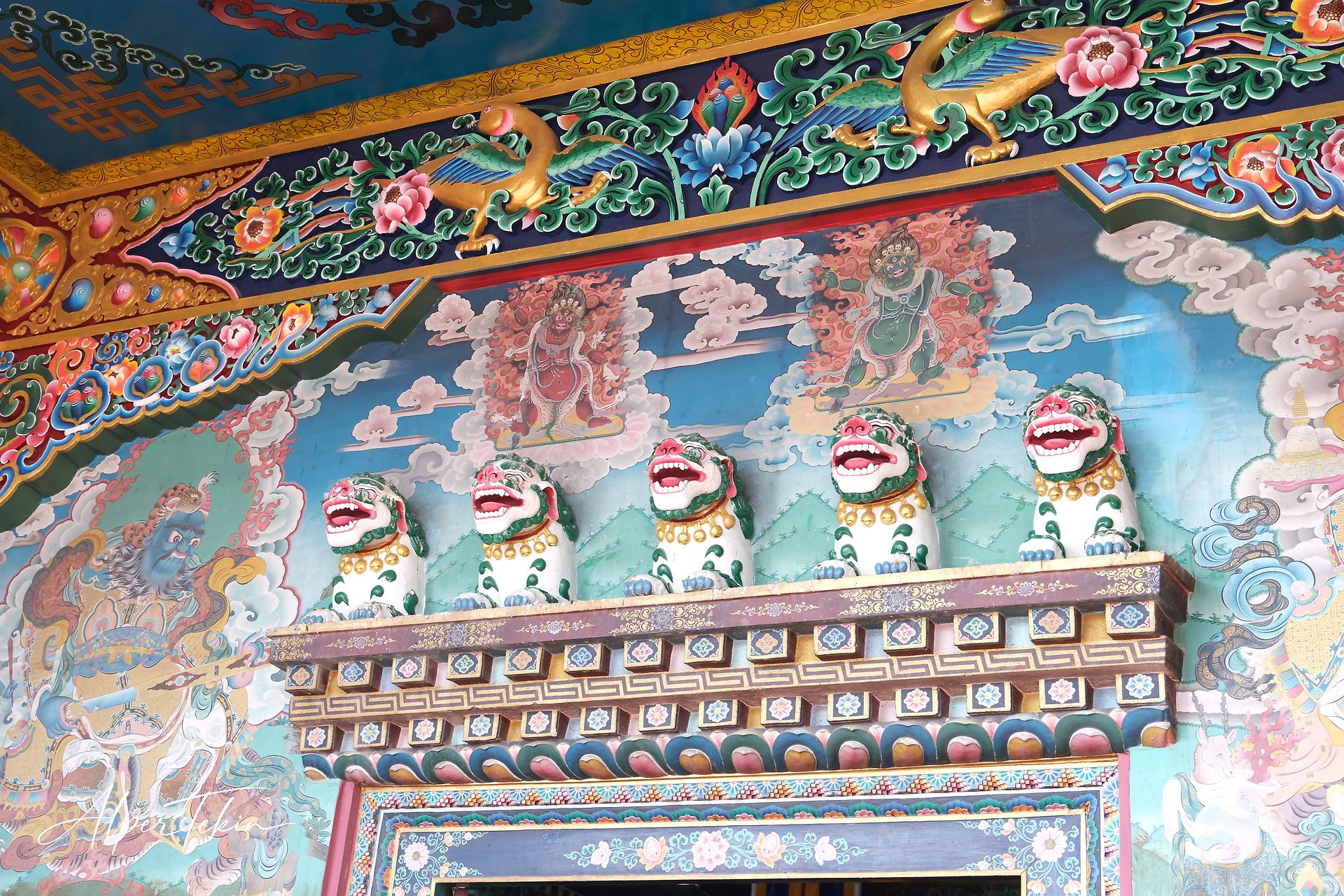 Five white and green snow lion figures line a ledge on a Tibetan-style entrance wall, with cloud murals behind and a gilded bird and floral frieze above.