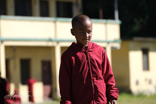 A shaven-headed boy in a red hooded jacket glances sideways, with a blurred yellow two-storey building and a patch of grass behind him.