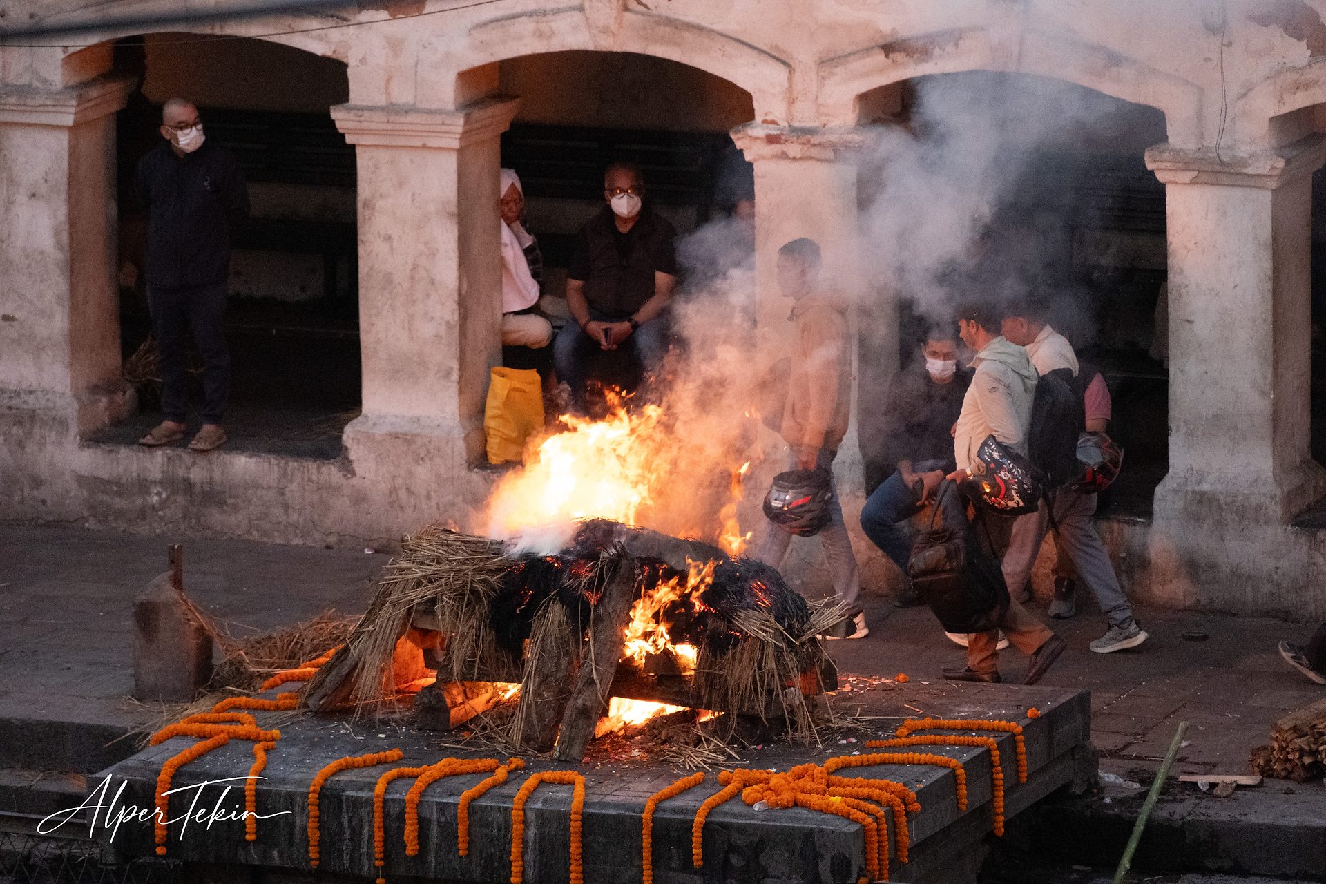 A burning pyre of wood and straw on a stone platform, edged with orange marigold garlands, with masked people and rising smoke beneath arches behind.