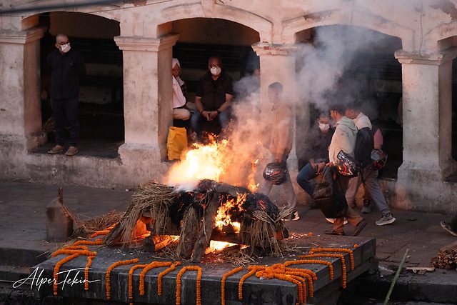 A burning pyre of wood and straw on a stone platform, edged with orange marigold garlands, with masked people and rising smoke beneath arches behind.