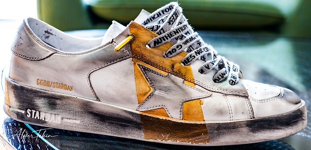 Side view of a distressed white leather sneaker with a gold star, printed laces and STARDAN lettering, resting on dark glass against a blurred green background.