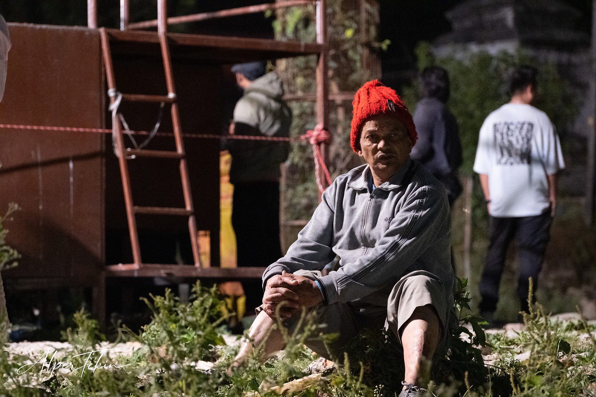 An older man in a red knit beanie and grey zip jacket sits among weeds at night, with a rusty scaffold and two blurred figures behind him.