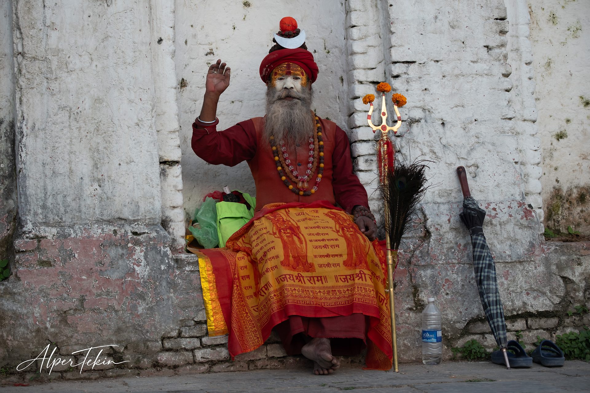 A bearded sadhu in a red turban and painted face sits before a weathered white wall, orange inscribed cloth across his lap, a flower-tipped trident beside him.