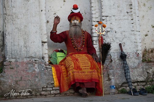 A bearded sadhu in a red turban and painted face sits before a weathered white wall, orange inscribed cloth across his lap, a flower-tipped trident beside him.