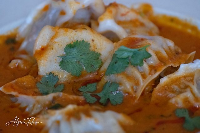 Close-up of folded dumplings in a orange-red sauce, garnished with green coriander leaves, with a shallow depth of field.