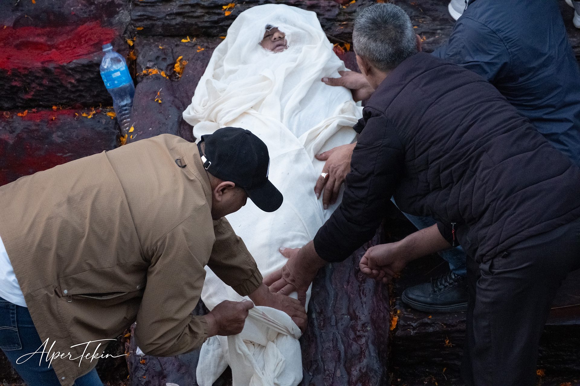 A person wrapped in white cloth with only the face uncovered, two men leaning over, on dark stone strewn with orange petals and red powder.
