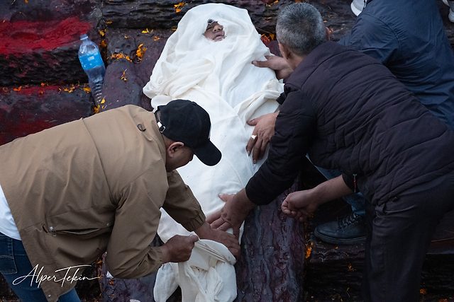 A person wrapped in white cloth with only the face uncovered, two men leaning over, on dark stone strewn with orange petals and red powder.