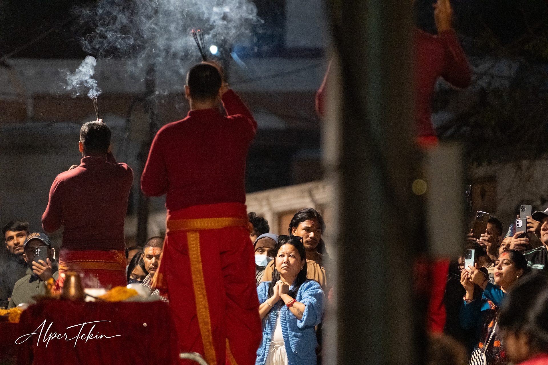 At night, two priests in red robes face away holding incense; in front of them a woman in a blue cardigan clasps her hands, with a phone-holding crowd around.