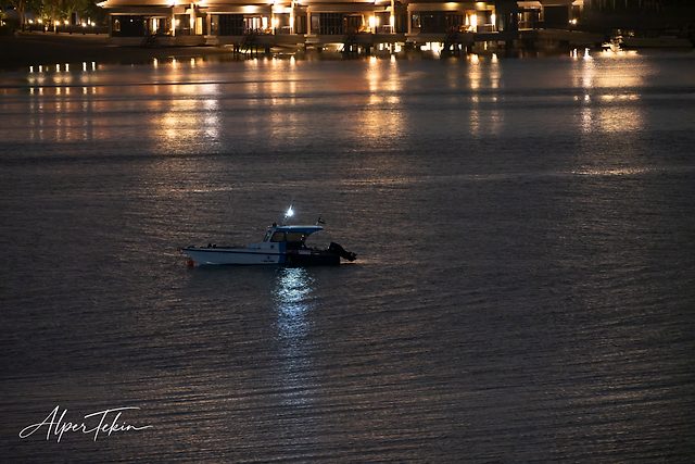At night, a white and blue boat sits on dark rippled water, with lit overwater buildings and orange reflections behind it.