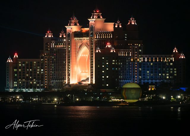 A large towered hotel lit in orange against the night sky, dark water in the foreground and a green and yellow lit balloon at right.