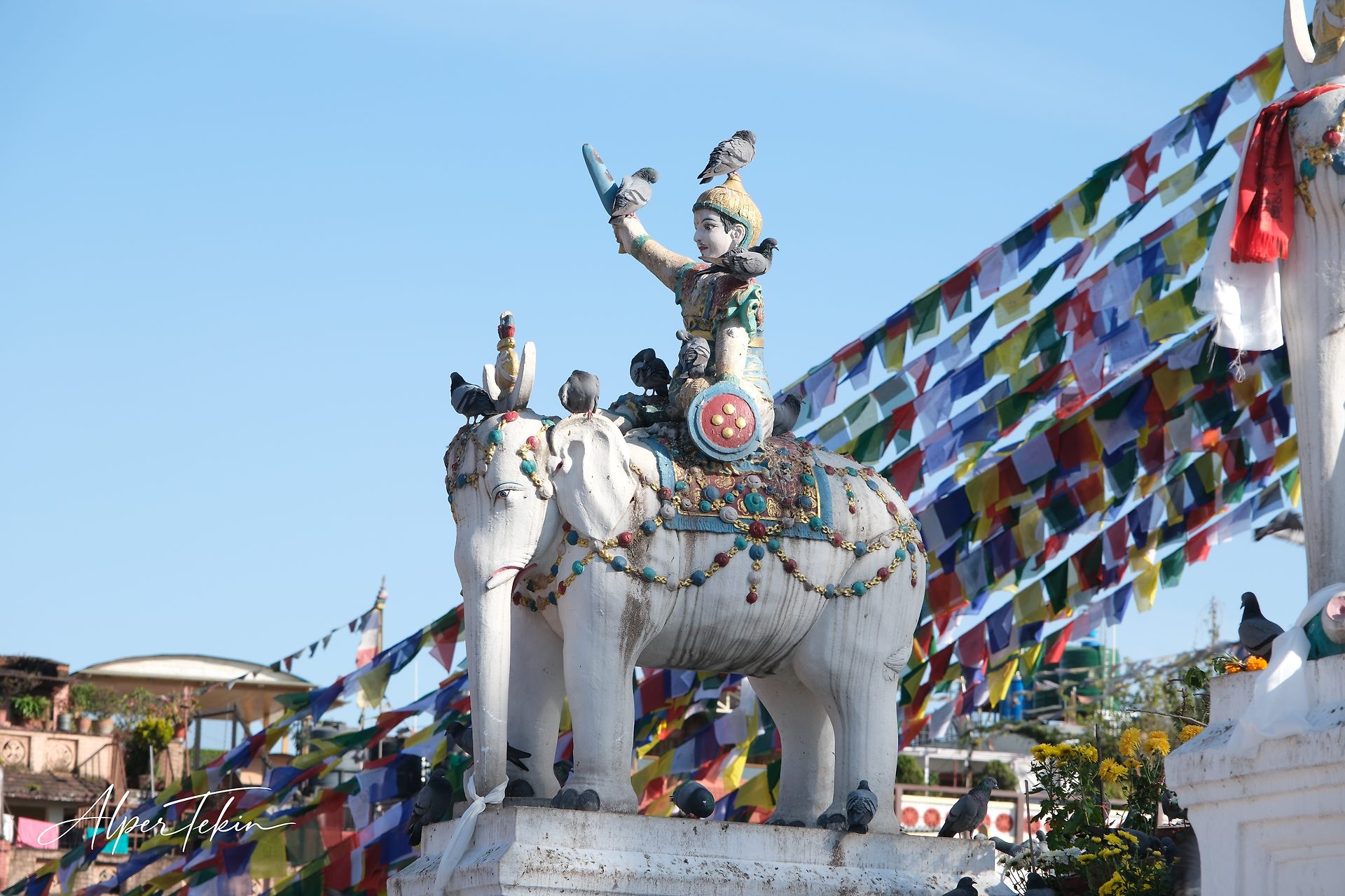 A painted figure rides a white elephant statue dotted with pigeons, set against a clear blue sky and diagonal strings of multicolored prayer flags.