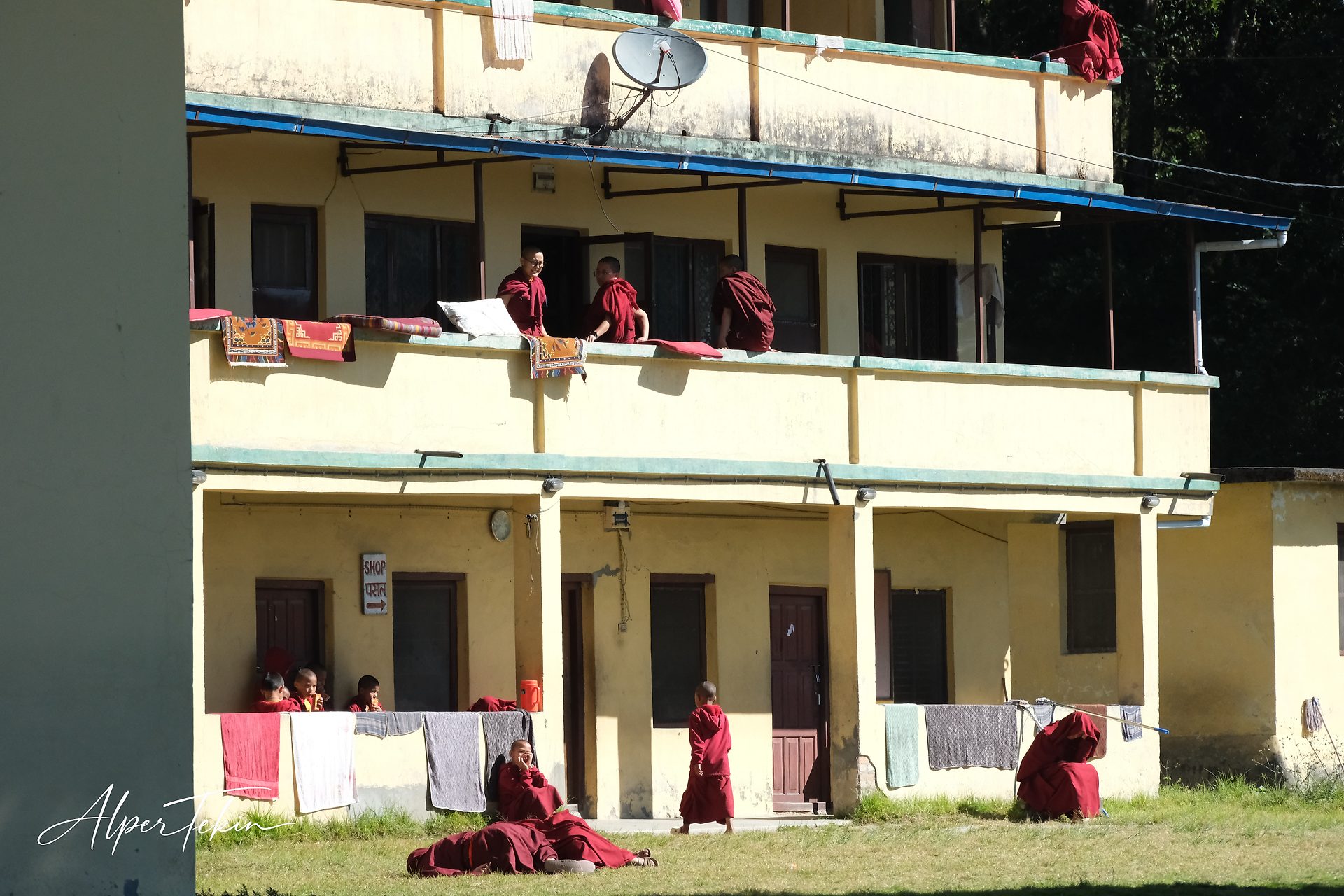 Monks and children in maroon robes on the balconies and lawn of a yellow three-story building, with rugs and laundry hung along the railings.