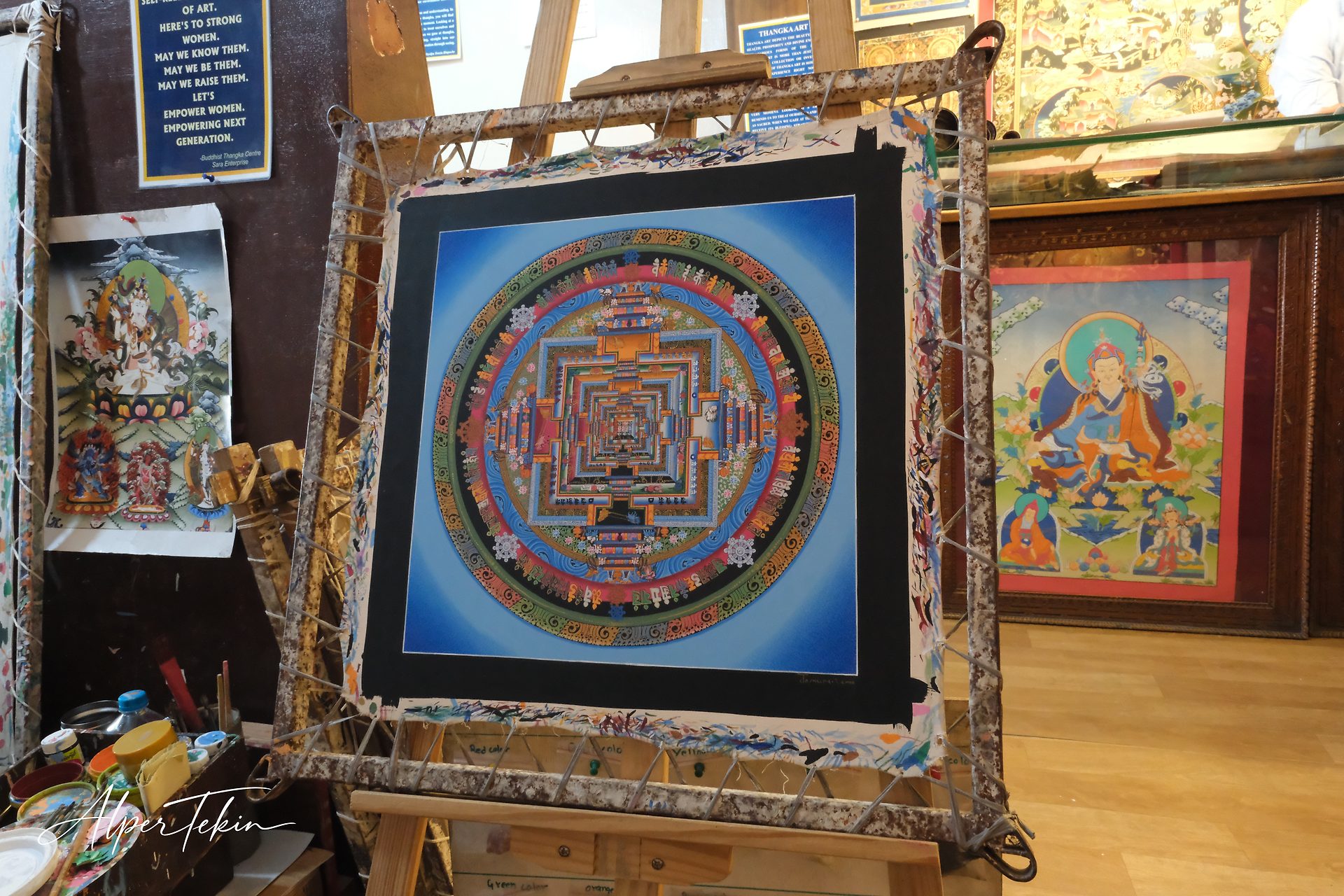 A blue thangka mandala with a black border, lashed to a wooden frame; other thangka paintings and paint pots are visible in the workshop behind.