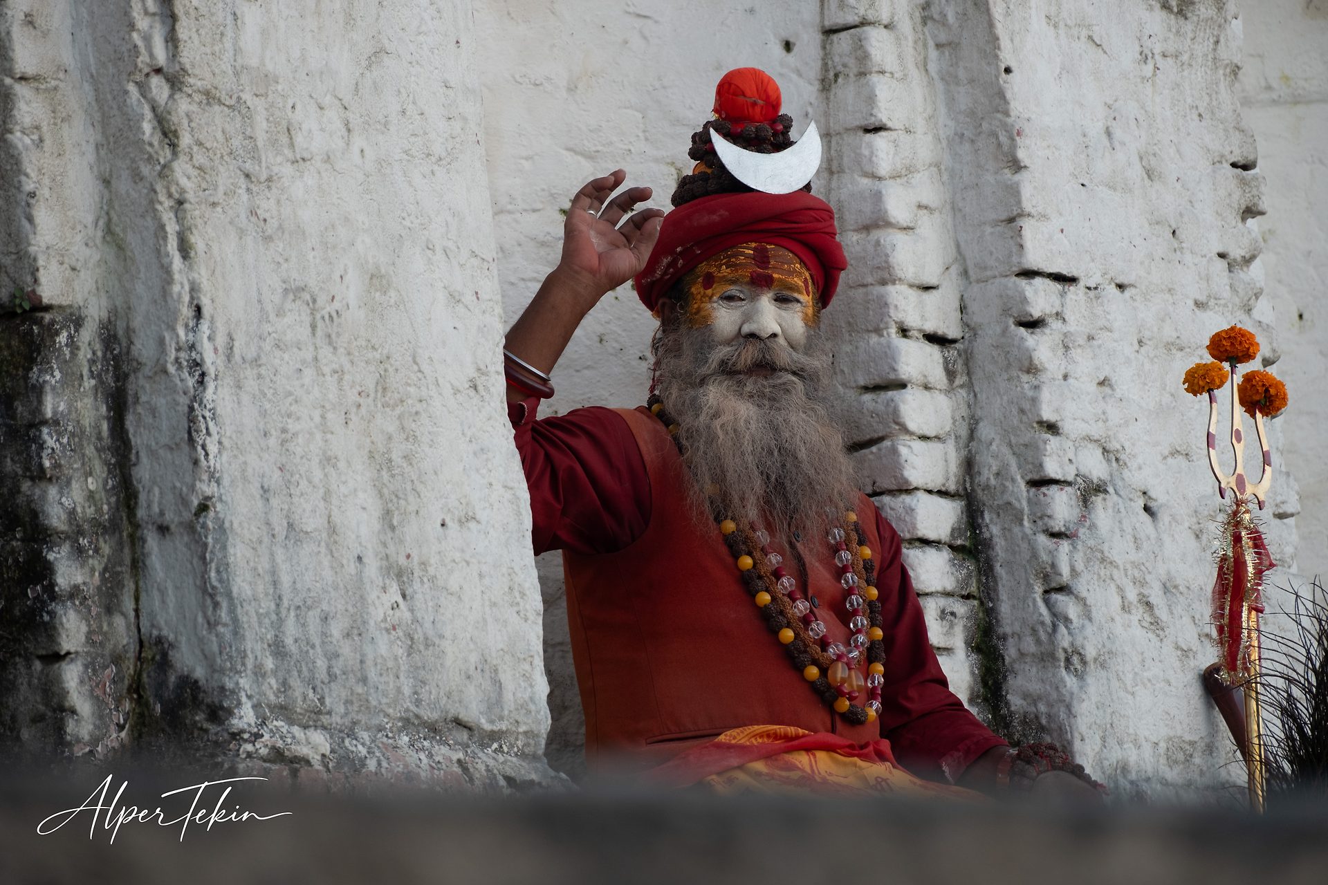 A long-bearded sadhu in a red turban and painted face sits with one hand raised, beads around his neck, a marigold-topped trident beside him.